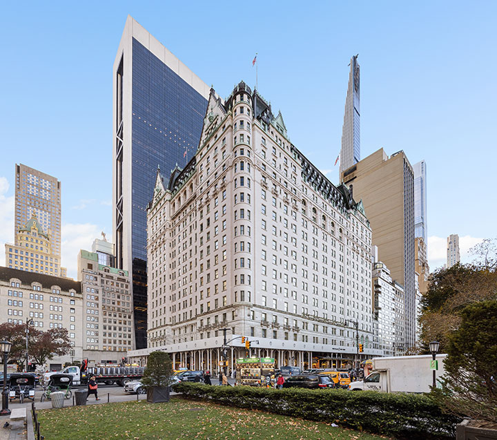 Exterior view of iconic New York City Plaza Hotel, nestled between skyscrapers and commercial buildings on a fall day with blue skies.